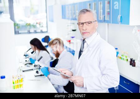 Travaux de laboratoire de trois. Gros plan portrait de professeur en costume formel et manteau de travail spécial dans les lunettes tenant la tablette et regardant sérieux et Banque D'Images