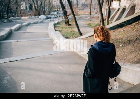 Vue arrière de la jeune femme vêtue d'un manteau noir et d'un sac à dos avec des cheveux brun frisé passe le long de la rue de la ville le jour ensoleillé du printemps. Banque D'Images