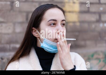 Une fille dans un masque antiviral prend et fume une cigarette dans la rue. Banque D'Images