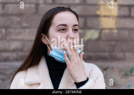 Une fille dans un masque antiviral prend et fume une cigarette dans la rue. Banque D'Images