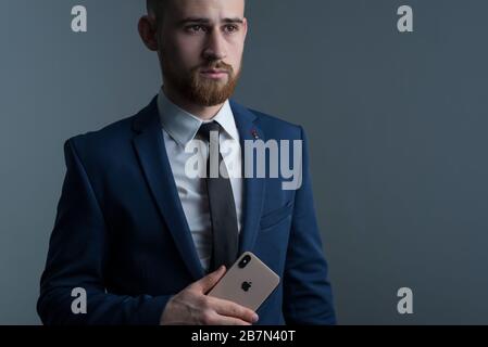 Zaporozhye. Ukraine. Février 2020. Gros plan portrait d'un jeune homme barbu dans un costume d'affaires tenant un iPhone XS dans sa main. Nouveau smartphone de Banque D'Images