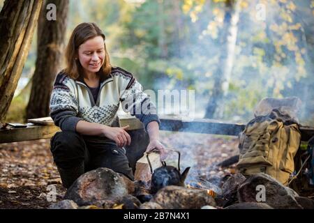 Homme et femme faisant du café dans la grande bouilloire sur feu de camp dans la forêt sur la rive du lac, faisant un feu, griller. Un couple heureux à explorer la Finlande. Scandinavie Banque D'Images