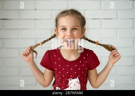 Adorable drôle fille tenant des cheveux de queue de porc sur un mur de brique. Petite fille mignonne avec de longs cheveux blonds à l'extérieur se défaire autour, montrant la langue Banque D'Images