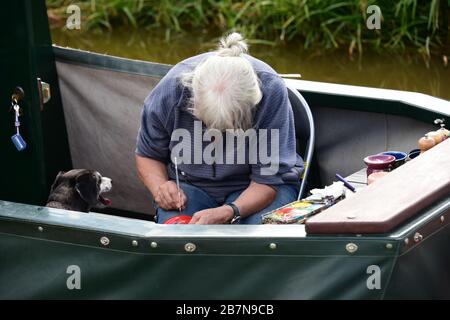 Femme aux cheveux gris peignant un article, regardé par son chien, sur la poupe d'un narrowboat pendant un festival de canal à Whitchurch, Shropshire Royaume-Uni Banque D'Images