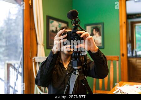 Vue de face et de gros plan avec mise au point peu profonde d'un homme caucasien assis derrière un appareil photo reflex numérique fixé sur un trépied avec microphone, filmant des vidéos à l'intérieur. Banque D'Images