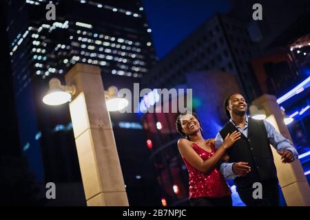 Happy young couple parler et rire comme ils marchent bras dessus bras dessous à travers la ville la nuit. Banque D'Images