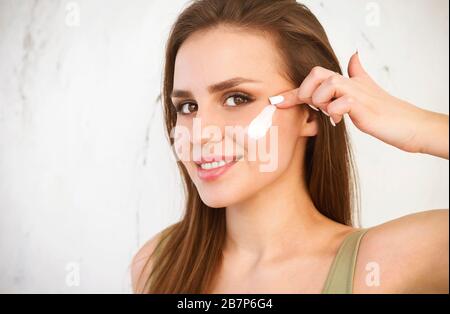 Jeune femme positive avec une belle peau frottant la crème cosmétique blanche sur la joue et regardant l'appareil photo tout en se tenant sur fond blanc Banque D'Images