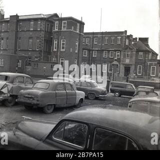 Années 1960, historique, voitures de l'époque, beaucoup d'entre eux ont endommagé et abandonné, garés sur un sol rugueux ou de déchets, en dehors d'un bâtiment hospitalier victorien, sud-est de Londres, Angleterre, Royaume-Uni. Banque D'Images