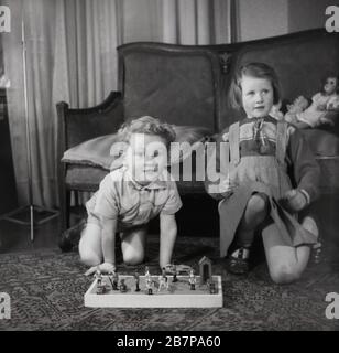 Dans les années 1950, historique, deux jeunes enfants dans une chambre d'entrée, une petite fille et un garçon près d'un canapé, peut-être frère et soeur, avec le garçon jouant avec ses soldats en jouet, Angleterre, Royaume-Uni. Banque D'Images