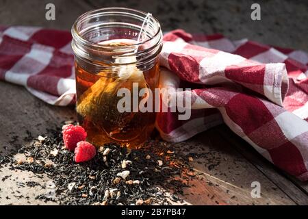 Sac à thé maison qui coule dans un pot de maçon, avec du thé et des framboises déversés devant, et un tissu à carreaux rouge et blanc derrière. Banque D'Images