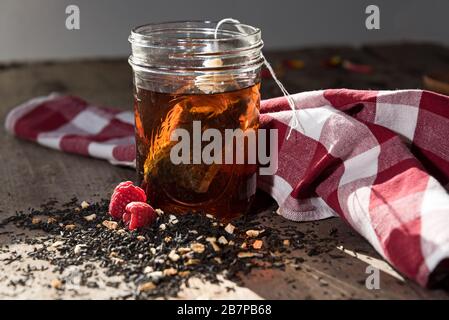 Sac à thé maison qui coule dans un pot de maçon, avec du thé et des framboises déversés devant, et un tissu à carreaux rouge et blanc derrière. Banque D'Images