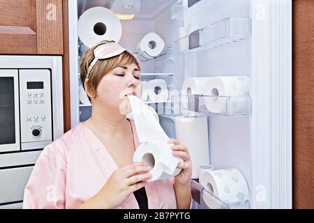 Une femme en pyjama mange du papier toilette près du réfrigérateur rempli de papier toilette. Concept de papier toilette sur-acheté pendant le coronavirus Banque D'Images