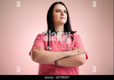 Portrait d'une femme magnifique médecin avec stéthoscope portant des gommages roses, regardant un héros-shot confiant posant sur un dos isolé rose. Banque D'Images