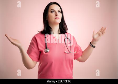 Portrait de belle femme médecin avec stéthoscope portant des gommages roses, avec des bras ouverts regardant vers le haut, vue latérale posant sur un dos isolé rose. Banque D'Images