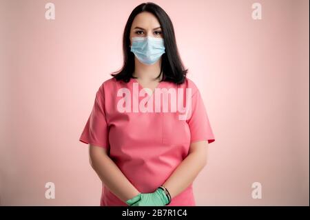 Portrait de belle femme médecin avec stéthoscope portant des gommages roses, avec masque de protection posant sur un dos isolé rose. Banque D'Images