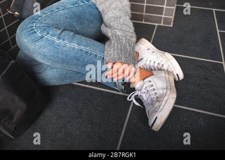 Gros plan image d'une femme portant des chaussures jean et blanches qui touchent sa jambe tout en étant assise sur le sol Banque D'Images