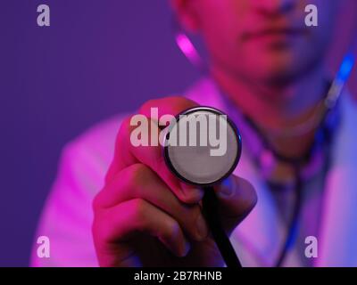 Portrait de héros en manteau blanc. Jeune médecin souriant et gai avec stéthoscope à l'hôpital médical, debout sur fond bleu et rose. Coronav Banque D'Images