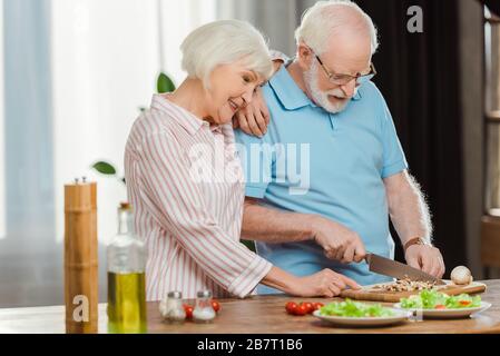 Femme senior souriante debout par mari coupant des légumes sur la table de cuisine Banque D'Images