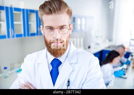 Gros plan portrait de jeune chercheur barbu rouge réussi, qui est en laboratoire, portant une tenue et un manteau formels, des lunettes Banque D'Images