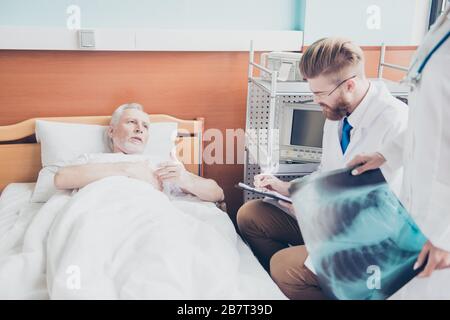 Le vieux patient dit à un jeune doc barbu rouge ses génomes du patient, il fait des notes, l'infirmière tient l'image de la radiographie Banque D'Images