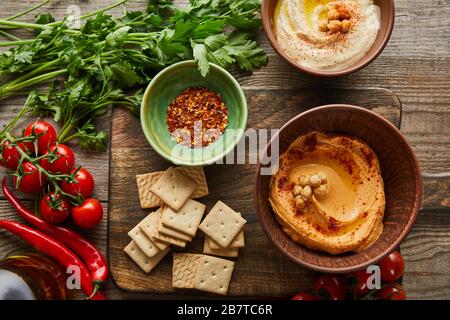 Vue de dessus des crackers, des bols aux épices et de l'houmous sur la planche à découper avec des légumes et du persil sur fond de bois Banque D'Images