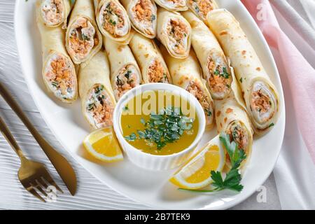 Crêpes farcies au fromage à la crème et au saumon, persil servi sur une plaque blanche sur une table en bois blanc avec sauce au beurre fondu, quartiers de citron, an Banque D'Images