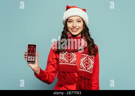 une jeune fille gaie à santa hat et un pull rouge montrant un smartphone avec un rythme de pulsation à l'écran sur fond bleu Banque D'Images
