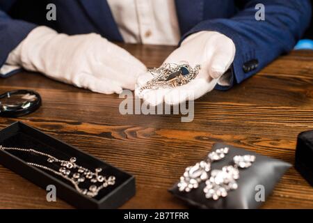 Vue rognée de l'évaluateur de bijoux tenant des bijoux près de la loupe sur la table sur fond noir Banque D'Images