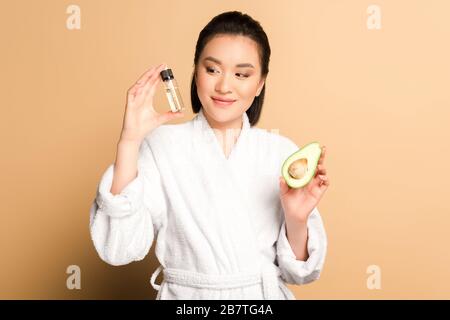 heureuse belle femme asiatique en peignoir avec avocat moitié et huile en bouteille sur fond beige Banque D'Images