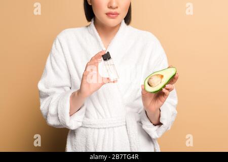 vue réduite d'une belle femme asiatique en peignoir avec moitié avocat et huile en bouteille sur fond beige Banque D'Images