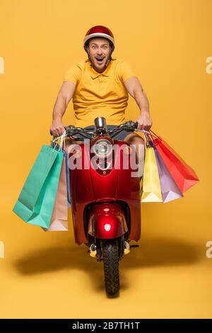 un homme de livraison heureux dans un scooter de conduite uniforme jaune avec des sacs de shopping sur fond jaune Banque D'Images