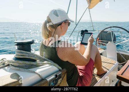 Femme assise sur le pont d'un yacht appréciant la lecture dans son ebook. Concept de voyage et de vacances. Banque D'Images