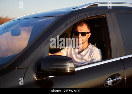 Un homme sérieux et confiant avec des poils dans un T-shirt blanc est assis dans sa voiture noire avec des lunettes de soleil sombres, regardant par la fenêtre de l'appareil photo, vue latérale, gros plan Banque D'Images