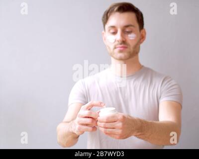 Jeune homme prenant soin de sa peau avec une crème d'hydratation et un closeup de lotion, fond. Banque D'Images