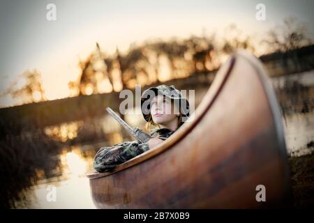 Jeune femme adulte assise en canoë avec une arme à feu près d'un lac. Banque D'Images