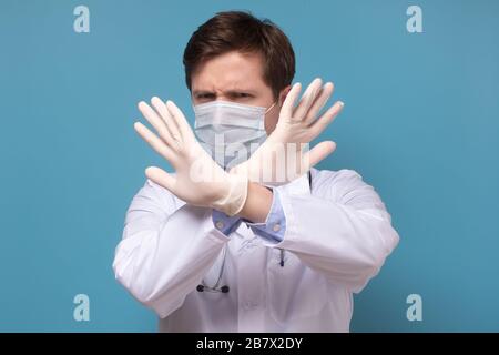 Beau jeune homme de médecin sur fond bleu isolé. Expression de rejet traversant les bras faisant signe négatif se protégeant. Banque D'Images