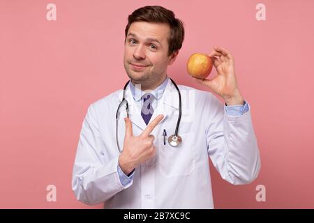 Un jeune médecin caucasien qui tient de la pomme fraîche qui l'indique. Concept de régime alimentaire sain. Studio tourné sur un mur rose Banque D'Images