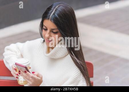 Une jeune fille moderne souriante discutant sur son téléphone mobile avec une tasse de café assis sur un banc dans la rue Banque D'Images