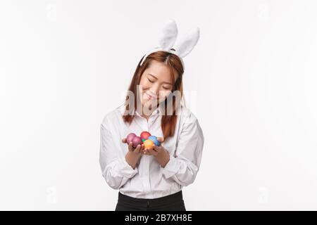 Vacances de Pâques, concept de célébration. Portrait d'une jeune fille asiatique timide et mignonne dans les oreilles de lapin, tenant des œufs peints et souriant, modeste Banque D'Images