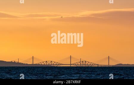 Firth of Forth, Écosse, Royaume-Uni. 18 mars 2020. Royaume-Uni Météo : un ciel orange coloré au coucher du soleil au-dessus du contour distinctif des trois ponts Forth avec un avion dans le ciel en descente à l'aéroport d'Edimbourg, vu de East Lothian Banque D'Images