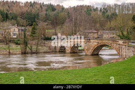 Un vieux pont en pierre s'étend sur une rivière au Prieuré de Kirkham dans le Yorkshire. Un bois est sur la colline au-delà et un ciel nuageux est au-dessus. Banque D'Images