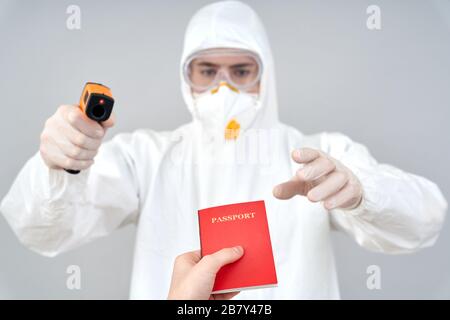 Concept de coronavirus. Médecin en costume de noisette et respirateur debout isolé sur gris prenant le passeport de la personne chinoise tenant la caméra infrarouge Banque D'Images