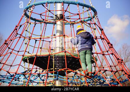 POZNAN, POLOGNE - 08 mars 2020: Un garçon portant des vêtements chauds grimpant sur un filet coloré d'un équipement dans une aire de jeux du parc Rataje. Banque D'Images