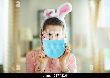 femme moderne dans un chemisier rose et des oreilles de lapin de pâques dans le salon moderne en journée ensoleillée de printemps se cachant derrière la plaque bleue en forme de poulet pour les œufs. Banque D'Images