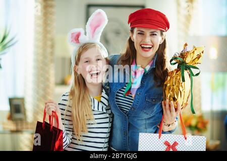 Portrait de la mère et de la fille souriantes et élégantes dans la maison moderne en journée de printemps ensoleillée tenant enveloppé dans de gros œufs de pâques en feuille d'or et sacs à provisions. Banque D'Images