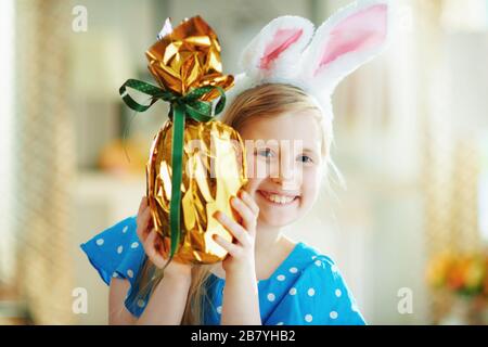 Portrait d'une heureuse petite princesse moderne dans un ensemble de pois bleus et des oreilles de lapin de pâques dans la maison moderne en journée ensoleillée de printemps regardant de wr Banque D'Images