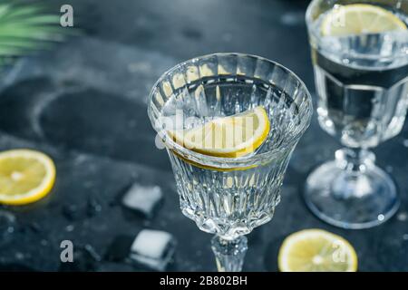 Détox de citron d'eau. Eau froide pure avec citron dans des verres en cristal sur un fond sombre dans le soleil ensoleillé d'été. Banque D'Images