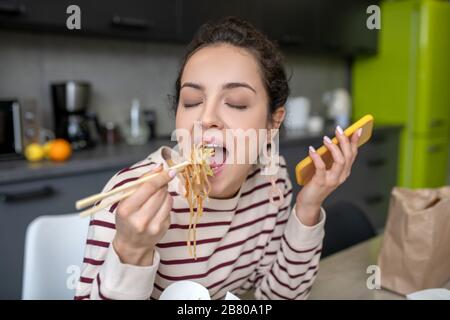 Jeune femme qui aime les nouilles dans la cuisine, tenant mobile Banque D'Images