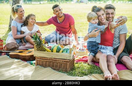 Les familles heureuses font un pique-nique dans le parc naturel en plein air - les jeunes parents s'amusent avec les enfants en été en prenant un selfie avec smartphone - technologie Banque D'Images