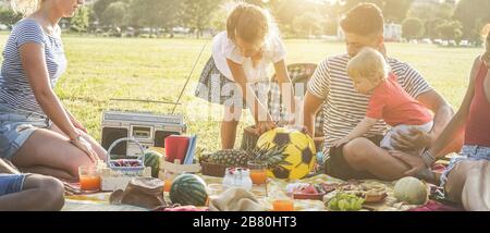 Les familles heureuses qui font un pique-nique dans le parc de la ville - les jeunes parents s'amusent avec leurs enfants en été en mangeant, en buvant et en riant ensemble - Love A. Banque D'Images